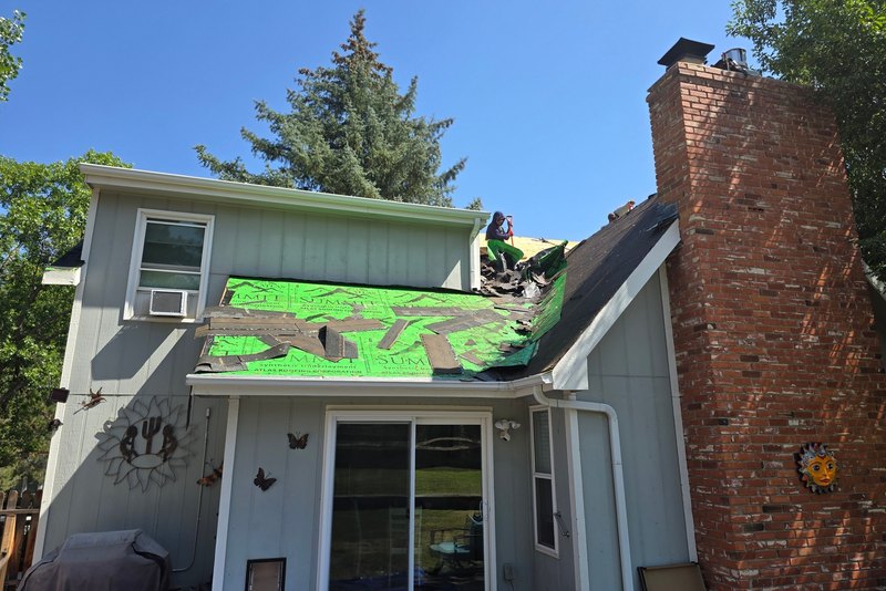 Partially stripped roof with green sheet material beside a brick chimney